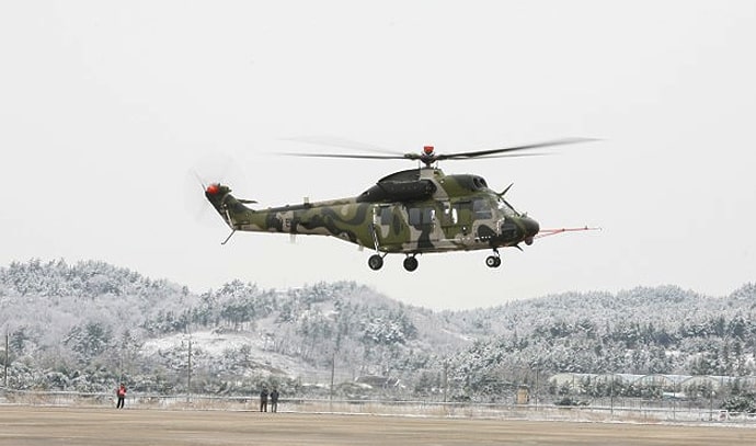 눈 덮인 풍경 위를 비행 중인 위장무늬 한국 육군 헬리콥터, 지상에는 작은 사람들이 서 있음