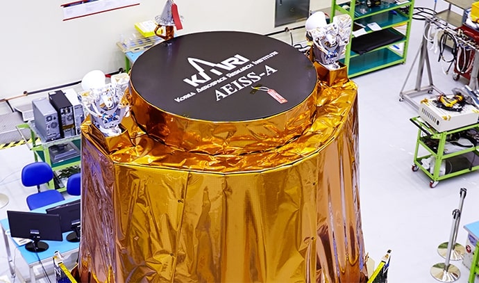 한국항공우주연구원(KARI) 마크와 ‘AEISS-A’ 라벨이 선명히 보이는 원형 덮개가 위에 설치된 위성 본체. 금색 방열재로 감싸져 있으며, 청정실 내부에서 점검 대기 중인 모습.