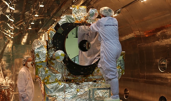 열진공 챔버 내부에서 은색 열차폐재로 둘러싸인 위성을 작업자가 사다리에 올라 점검하고 있는 모습. 작업자는 흰색 방진복과 마스크를 착용하고 있으며, 주변은 금속 내부 벽으로 둘러싸여 있음.