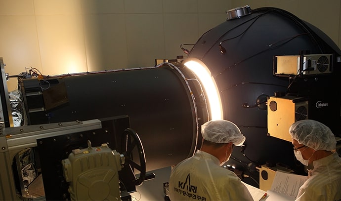 광학 시험 장비에 연결된 위성 부품에서 밝은 빛이 나오는 상태에서 두 명의 연구원이 도면을 보며 검토하는 모습. 연구원들은 KARI 로고가 새겨진 방진복과 모자를 착용하고 있음.