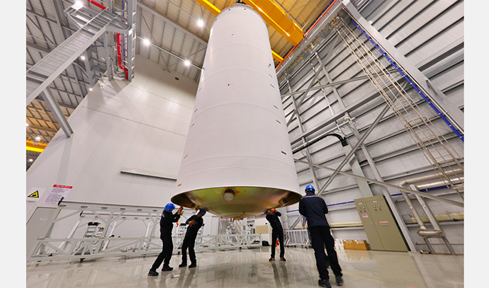 크레인에 매달린 대형 발사체 탱크를 명의 작업자가 바닥에서 손으로 받쳐가며 수평을 맞추는 장면. 내부는 넓은 조립동이며 벽에는 구조물과 배선이 보임.