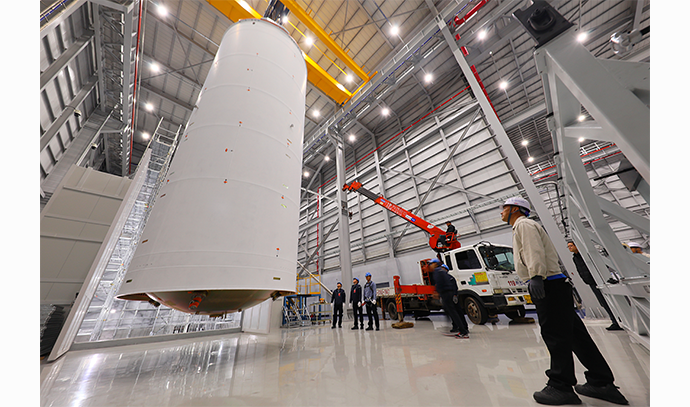 크레인에 매달린 대형 발사체 탱크가 기울어진 상태로 이동 중이며, 작업자들이 주위를 지켜보고 있는 모습. 옆에는 소형 크레인이 대기 중임.