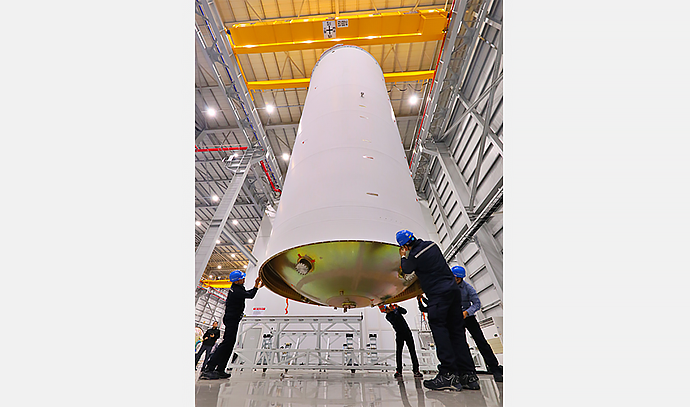 다섯 명의 작업자가 대형 발사체 탱크의 하부를 받쳐 수평을 맞추는 장면을 다른 각도에서 촬영. 높은 천장의 대형 실내 공간과 노란색 크레인이 배경에 보임.