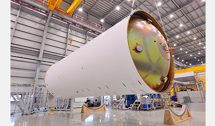 초대형 발사체 탱크가 특수 받침대에 수평으로 거치된 상태. 주변에는 조립 설비와 계단, 작업대가 있으며 공장 내부가 밝게 조명되어 있음.