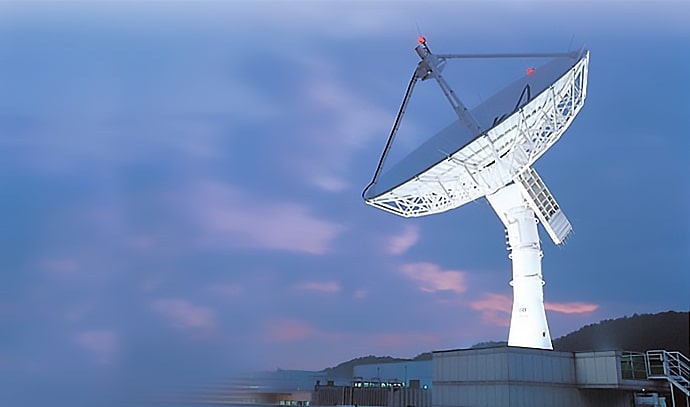 해 질 녘 보랏빛 하늘을 배경으로 지상국 옥상에 설치된 대형 흰색 패러볼라 안테나가 하늘을 향해 기울어져 있다. 안테나 거더와 상단에는 붉은 항공장애등이 켜져 있다.