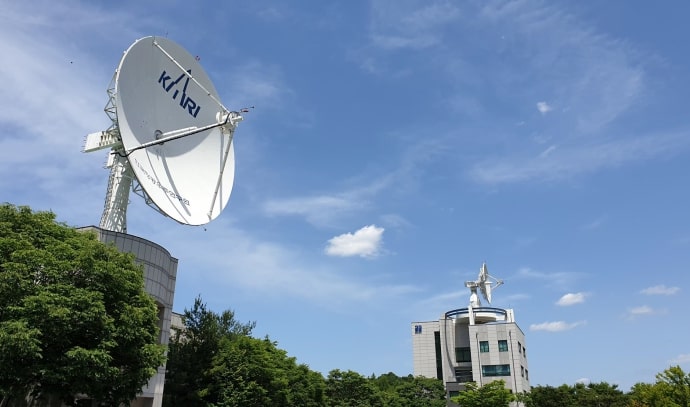 맑은 낮 하늘 아래, 왼쪽에는 KARI 로고가 그려진 거대한 흰색 위성 안테나, 오른쪽에는 별 모양 조형물이 얹힌 연구동 건물이 보인다. 전경에 푸른 나무와 하늘에 흰 구름이 배경을 이룬다.