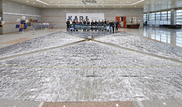 한국항공우주연구원(KARI) 로비에 설치된 10m 크기의 태양돛(Solar Sail) 전개장치를 전시한 사진. 돛 중앙에는 장치를 조작한 연구진들이 단체로 서 있으며, '태양돛 전개장치 시험을 기념합니다'라는 현수막이 뒤에 걸려 있다. 얇고 반짝이는 재질의 정사각형 돛이 바닥에 넓게 펼쳐져 있다.