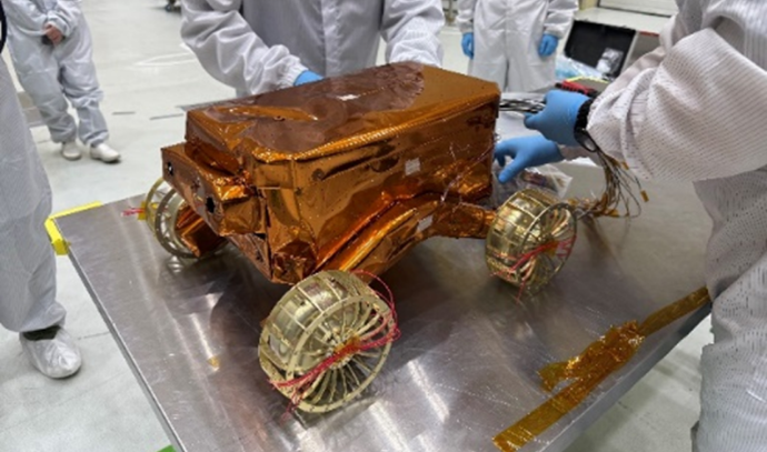 금색 열차폐 필름으로 덮인 소형 월면 탐사로버가 실험대 위에 올려져 있으며, 연구원들이 방진복과 장갑을 착용한 채 장치를 조작하고 있다. 탐사로버는 4개의 바퀴가 달린 직육면체 구조를 갖고 있다.