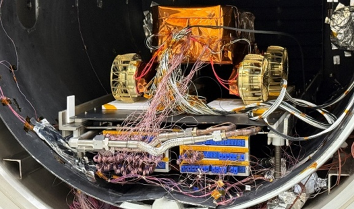 복잡한 전선이 연결된 상태로 진공 챔버 안에서 테스트 중인 소형 탐사로버. 위쪽에는 금박으로 덮인 본체가 있고, 아래쪽에는 각종 센서와 케이블이 연결된 제어 시스템이 장착되어 있다.