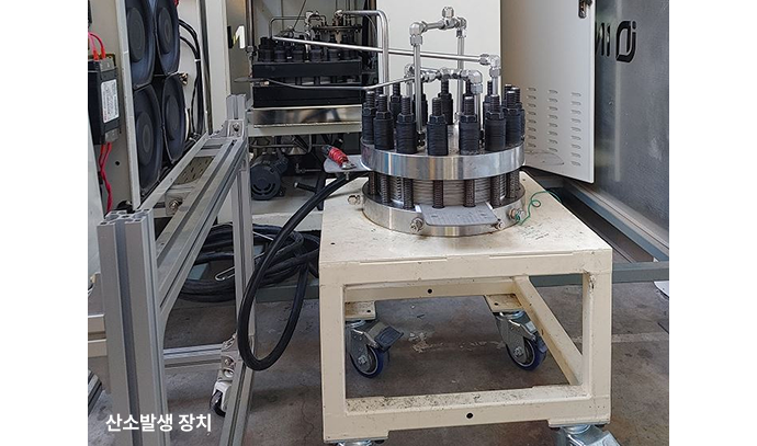 산소발생 장치의 실물 이미지. 바퀴가 달린 받침대 위에 원형의 PEM 전해조가 설치되어 있으며, 상단에는 다수의 전극과 케이블이 연결되어 있다. 주변에는 공조 장치와 전력장비가 배치되어 있다.